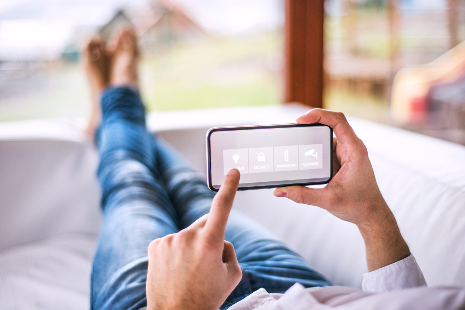 Otthonában pihenő férfi kezében okostelefonjával applikáción keresztül irányítja okosotthonának világítás funkcióját.